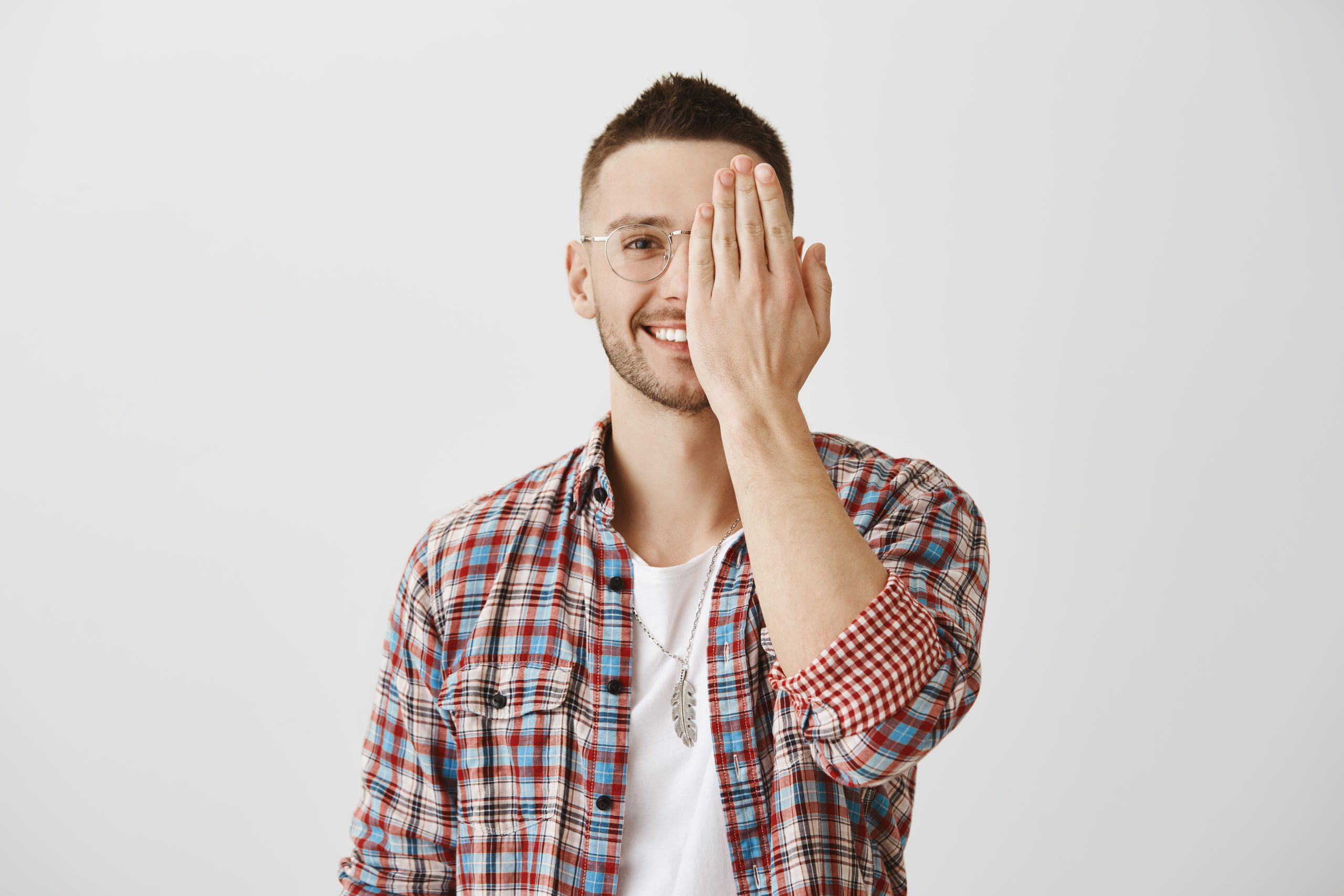 people emotions concept. portrait of attractive man in glasses hiding half of face with palm while smiling and expressing friendliness. guy is showing only positive side of his temper